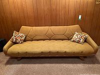 Full front view of mustard-yellow upholstered sofa on wooden frame with two floral accent cushions against wood-paneled wall.