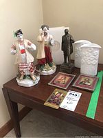 Porcelain figurines, two white porcelain vases, two small framed religious icon prints, and metal figurine on a table corner.