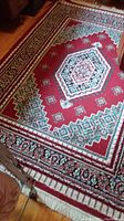 Top view of red decorative rug with green and white geometric and floral patterns on wooden floor with fringed edges visible.