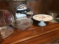 Silverplated oval platter, pinwheel crystal cake stand, Limoges porcelain footed cake stand, and other clear glass servingware on wooden cabinet surface.