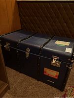 Photo of vintage navy blue steamer trunk with black straps, metal clasps, and travel stickers on the side.