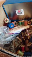Box with assorted spools of sewing thread, mostly wooden spools, visible threads in blue, green, orange, and red hues, a metal tin and some plastic wrapped bundles.