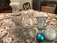 Arrangement of six items including two clear glass vases, a square ceramic vase with a label and pink trim, a beige ceramic vase with ornate details, a blue ornament ball, and a spherical vase filled with feather-like material.