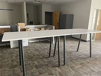 Photo shows a long drafting table in an office space with a rectangular light-colored top and dark metal legs standing on carpeted floor.