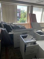 Photo showing multiple white metal file cabinets with open drawers, a stack of unassembled gray partitions, and desk pieces stacked near large windows.