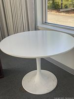 White round table with a single pedestal base, placed near window and curtain, showing surface and base condition.