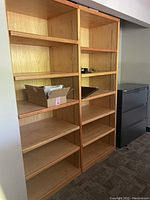 Light-colored wooden shelving units with multiple adjustable shelves, containing a cardboard box and some miscellaneous items on one shelf.