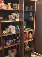 Wooden kitchen cabinet shelves with multiple boxed and packaged kitchen supplies including light bulbs, plastic wrap, foil, paper towel, and bottles.