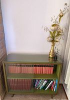 Full front photo of the green bookshelf holding a set of red books and assorted books beneath, along with a gold vase with artificial flowers on top.