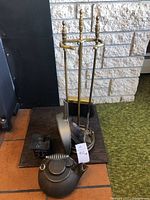 Front view of the brass fireplace tool set on circular base with attached brush and ash shovel, alongside cast iron matchbox and wood stove kettle.