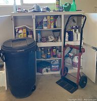 White cabinet open showing shelves with assorted lawn and cleaning supplies including spray bottles and chemical containers, with black plastic garbage bin and red hand truck in front.
