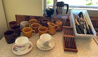 Photo showing multiple coffee and tea cups, plastic container with silverware, a wooden utensil holder, and a coffee press on countertop
