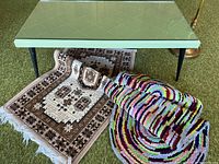 Front angled view of coffee table with black legs and glass top, brass floor lamp's base visible, two rugs under table: a beige and brown geometric patterned rug and a multicolor looped rug