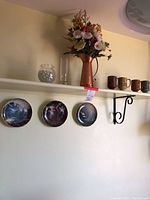 Wide view showing the three Franklin Mint collector plates, copper pitcher with flowers, small glass container, and pottery cups on a wall shelf.