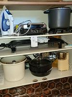 Wide shot showing shelves with various kitchen appliances and cookware including an iron, electric griddle, waffle maker, Crockpot, large pots, and a coffee/spice grinder