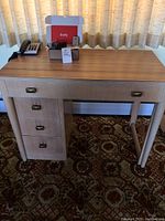 Full view of vintage wooden desk with three left-side drawers showing office items on top including a black corded telephone, lively alert device in box, playing cards, lanyard, and stapler.