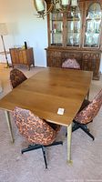 Long view showing wooden rectangular dining table with golden metallic legs and six chairs: four with orange floral fabric swivel bases and two floral chairs with wooden frames.