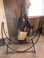Wide shot of metal firewood log holder with plastic and metal buckets filled with wood kindling and logs near window by wood-paneled wall.