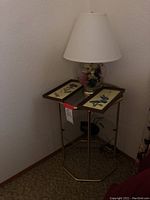 Full view of glass and brass nightstand with table lamp and two framed art pieces on top, placed in corner of room.
