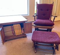 Gliding rocking chair with burgundy upholstered cushions, matching ottoman in front, and wooden end table with two doors containing books.