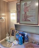 Two mirrored trays, cut glass soap dish, milk glass soap dish, assorted soaps, cleaning materials, and two framed, matted prints visible on sink counter and wall above.