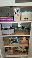 Overview of shelf with purple Sparkling Apple Grape box holding decorative Christmas ornaments, Premium Saltine Crackers tin, Brother label printer in box, paint brushes, and storage containers with miscellaneous items.