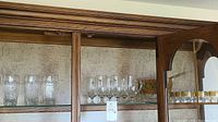Glassware including clear textured and smooth drinking glasses on the top glass shelf inside wooden cabinet.