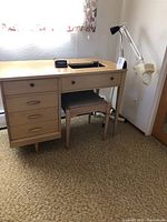 Retro sewing cabinet with drawers, attached small bendable lamp, upholstered stool below, Sharp alarm on top, and magnifying lamp to the right.