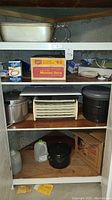 Kitchen shelving unit holding multiple kitchen items including large roasting pan, Mason jar caps in boxes, pots, squeezo strainer and other utensils.