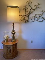 Full view of vintage lamp on octagonal end table with hurricane glass globe and metal leaf wall art on wall behind the lamp
