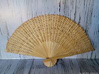 Light colored semi-circular wicker fan with detailed weaving on wooden slats, displayed on a weathered wood background.