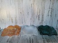 Three vintage Indiana Glass diamond point pattern ruffled edge bowls in marigold, milk glass with iridescent trim, and deep teal colors, displayed side by side against a wood plank background.