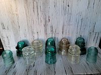 Photo shows lot of 10 vintage glass insulators in two rows, colors range from clear to dark turquoise green. Aged condition with chips noted on some items.