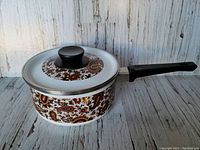Side view of the medium-sized enamel saucepan with brown paisley pattern and black handle against a white wooden background.
