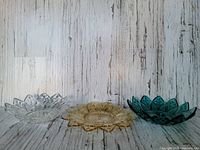 Front view of set of three floral shaped Federal Glass dishes in clear, amber, and deep teal colors, placed side by side on a wooden textured background.