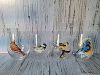 Front view of four stemless wine glasses with individual colorful bird designs: red cardinal, black-capped chickadee, yellow goldfinch, and bluebird, against a light wood background.