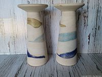 Pair of vintage thick pottery candlesticks with abstract colored bands on a wood floor and distressed wood backdrop, angled view showing height and decoration.