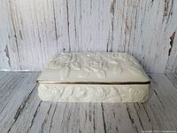 Ceramic jewellery box showing embossed rose design on all sides with gold trim on lid edge, placed on textured wood background.