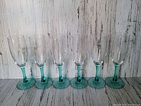 Front view of six aqua stemmed champagne flutes lined up side by side on a wooden surface with a white and gray distressed background.