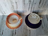 Two Aynsley teacup and saucer sets, one burnt orange with gold scroll, and one blue with gold leaf design, side by side showing top view