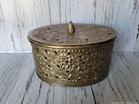 Front angled view of the vintage oval brass box showing intricate floral and botanical cutout patterns and pinecone-shaped finial on the lid.