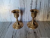 Two short brass candlesticks against a wood-patterned background, showing front and side angles with visible original patina.