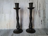 Pair of vintage heavily cast iron candlesticks with round bases and tapered shafts, shown side-by-side against a textured background.