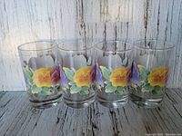 Set of four Corelle juice glasses with Summer Blush floral pattern arranged side by side on a rustic wood surface with a weathered white wood background.