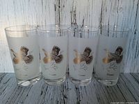 Four frosted Dominion glasses showing flying grouse bird design from front angle on light wooden background.