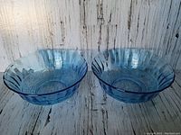 Two translucent ice blue glass bowls side by side on a rustic wood surface, showing scalloped edges and embossed design.