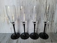 Four vintage Luminarc champagne flutes with black stems and clear bowls, shown standing in a row against a white rustic wooden background.