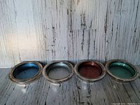Front view of set of four vintage silver deep coasters with blue, white, red, and green enamel interiors aligned in a row on a white wooden surface.