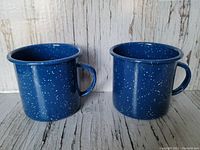 Front view of two vintage blue speckled enamel metal camping mugs showing their exterior and handles.