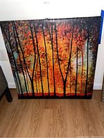 Full view of the framed artwork showing a forest with orange foliage and black tree trunks, laid on the floor against a wall.